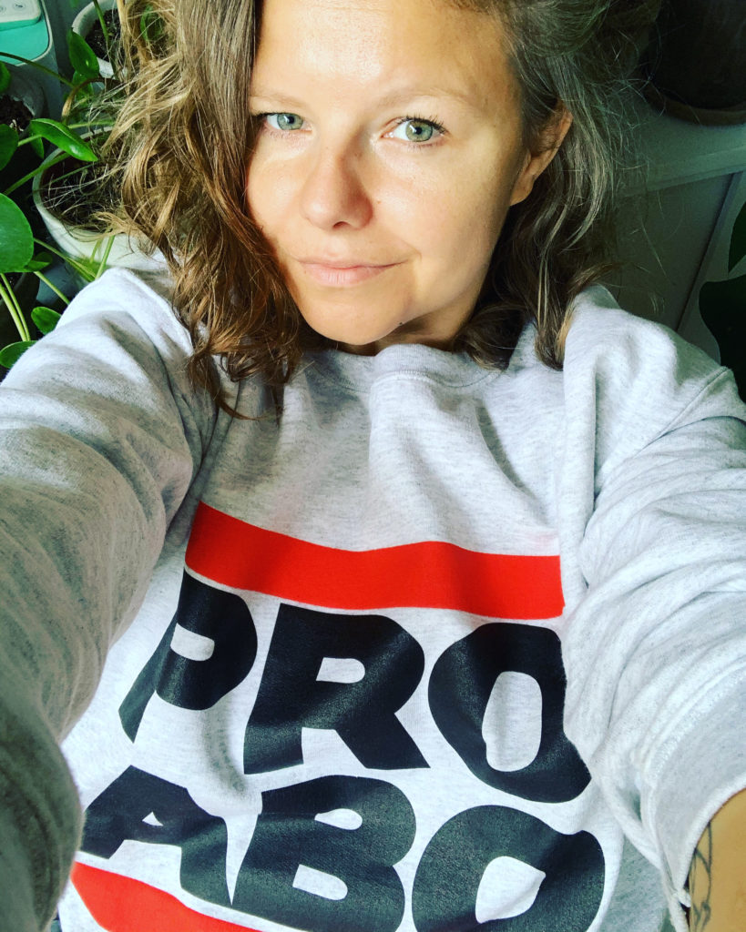 A person with green/grey eyes and knee length blonde hair, wearing a hoodie that says PRO ABO takes a selfie